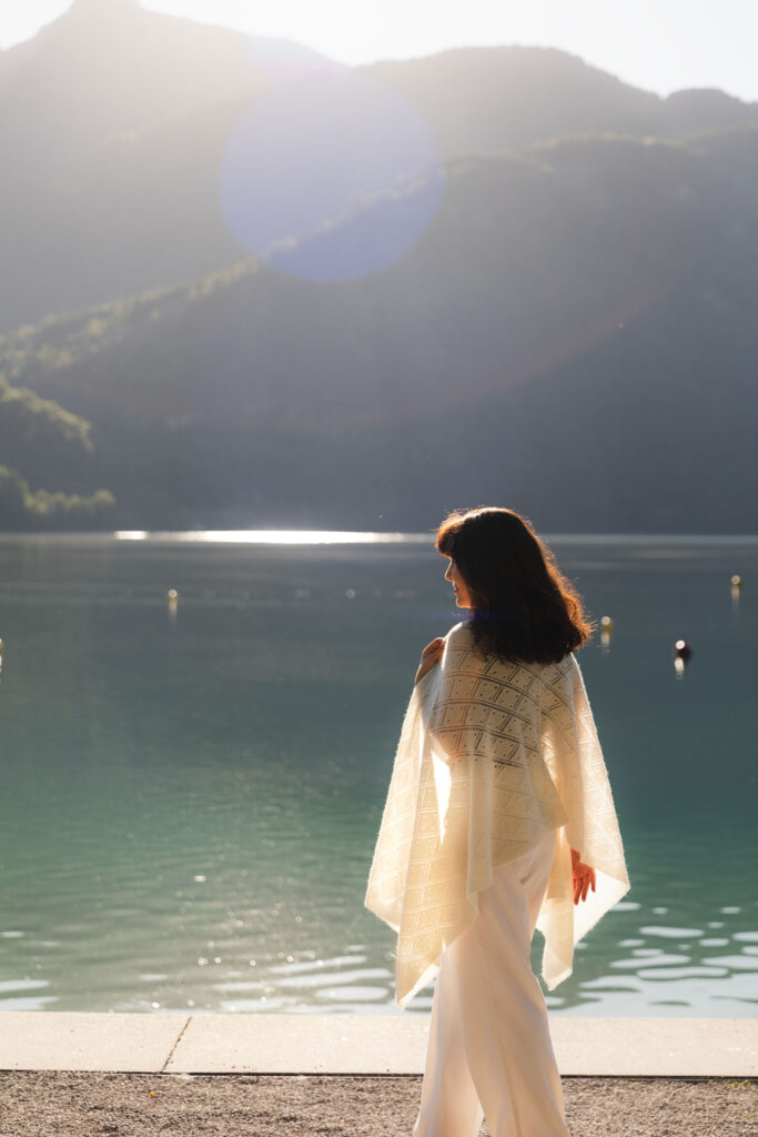 Woman wearing a white lace cashmere shawl while sitting beside a mountain lake at sunrise.