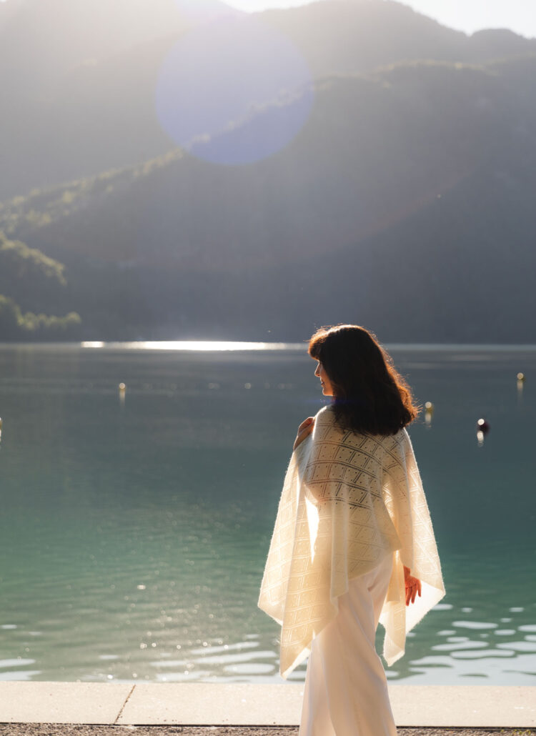 Woman wearing a white lace cashmere shawl while sitting beside a mountain lake at sunrise.